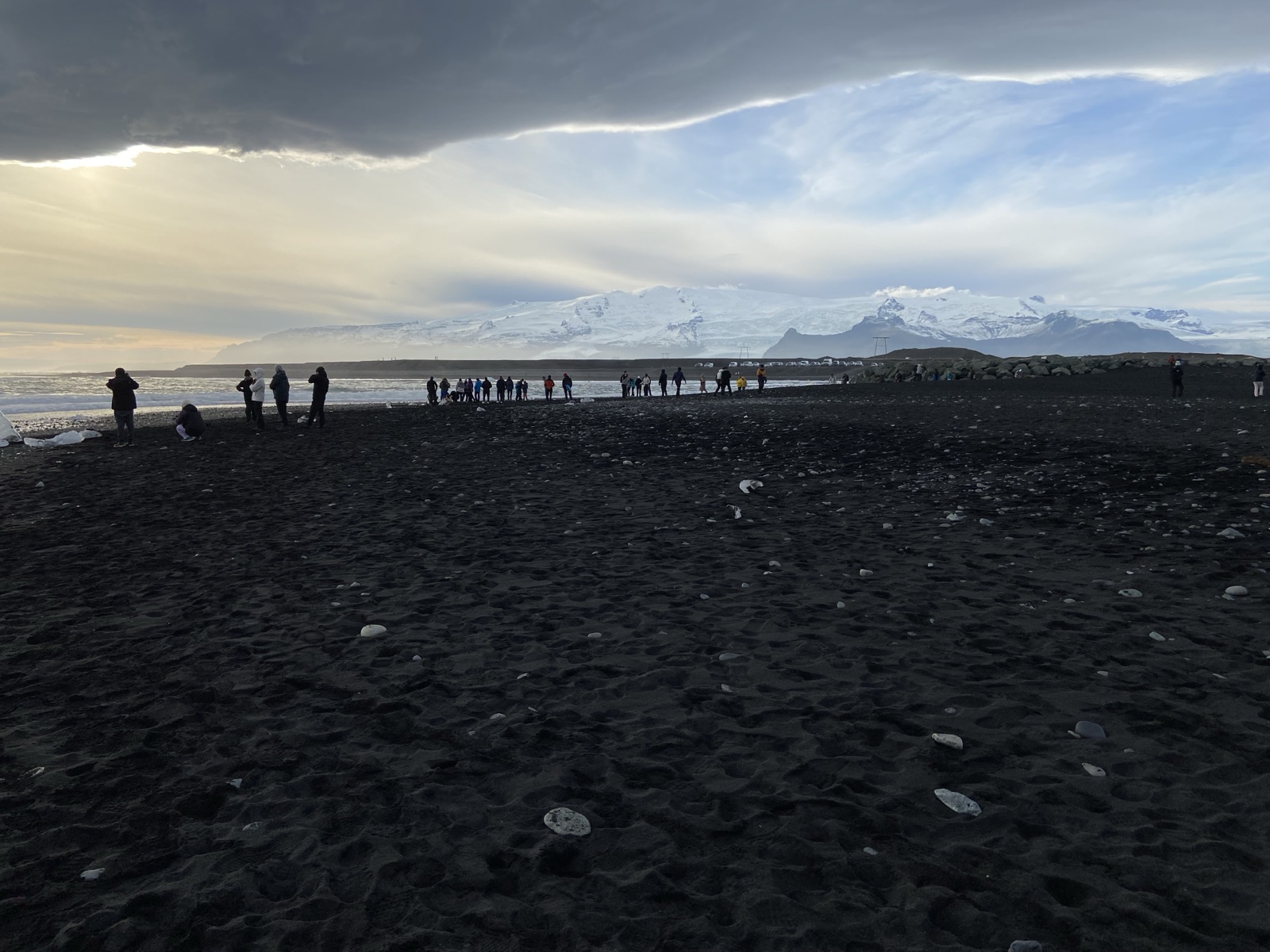 People gathered on a black-sand beach below snowcapped mountains at dusk