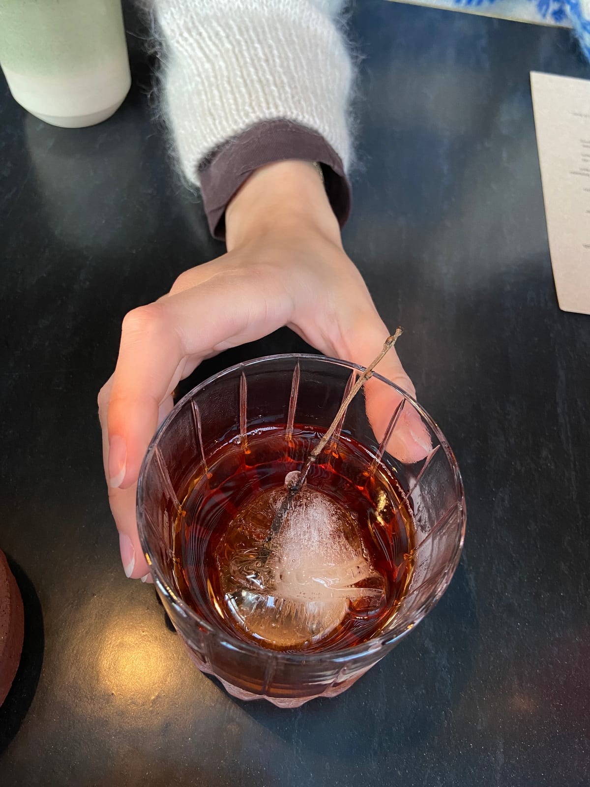 A hand around a cut-crystal glass of a dark cocktail over a clear ice ball, garnished with a sprig