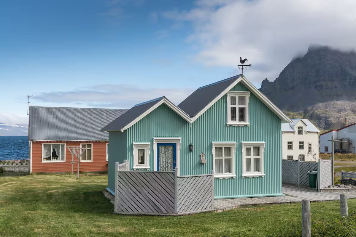A teal corrugated-iron cottage by the water below a fog-topped mountain in the Westfjords