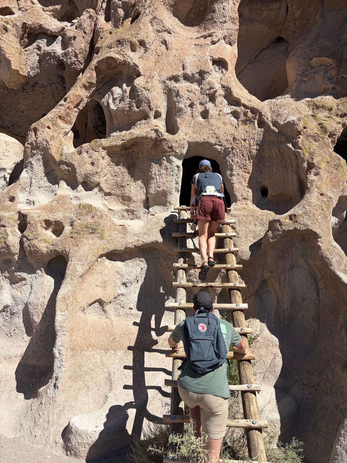Two visitors climbing a wooden ladder to a cave dwelling carved into the cliff at Bandelier