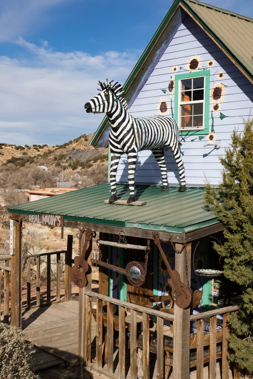 A metal zebra sculpture standing on the porch roof of a lilac clapboard gallery painted with sunflowers in Madrid, New Mexico