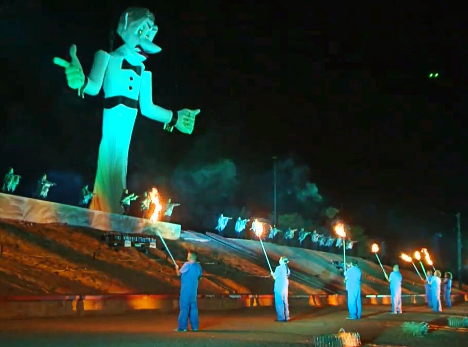 The fifty-foot Zozobra effigy lit green at night above a line of fire dancers holding torches